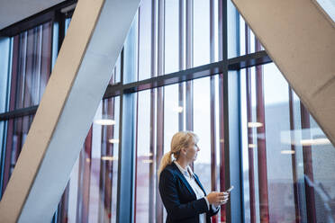 Blond businesswoman holding smart phone and looking through window - JOSEF23983
