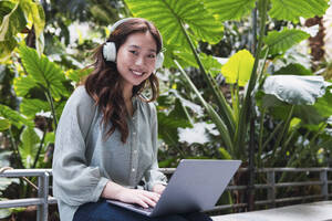 Lächelnde Frau mit Laptop und drahtlosen Kopfhörern auf einer Bank sitzend - PNAF06166