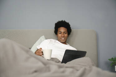 Germany, happy young man reading tablet, holding coffee in hand - FLMZF00008