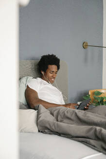 Germany, young man lying on bed, watching movie on a tablet - FLMZF00012