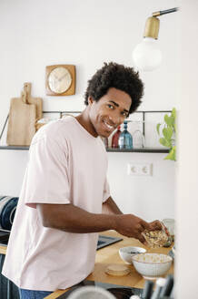 Smiling man preparing breakfast muesli in bowl at kitchen counter - FLMZF00029