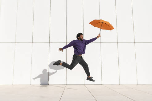 Fröhlicher Mann mit orangefarbenem Regenschirm vor einer weißen Wand springend - VRAF00476