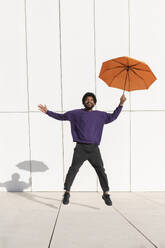 Portugal, young man jumping with orange umbrella against the white minimalistic tile wall - VRAF00477