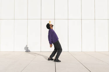 Young man balancing cup on forehead in front of white wall - VRAF00479