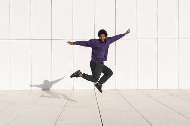 Playful young man jumping with arms outstretched in front of white wall - VRAF00481