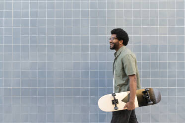 Happy man walking with skateboard in front of tiled wall - VRAF00485