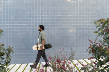 Young man walking with skateboard in front of blue tiled wall - VRAF00486
