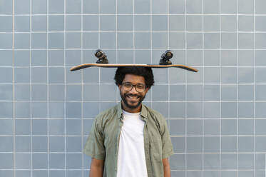 Portugal, young funny man holding a skateboard on his head against the blue tile wall - VRAF00489