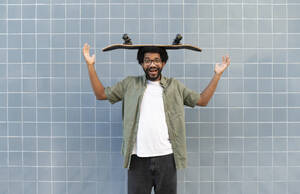 Playful man balancing skateboard on head in front of tiled wall - VRAF00490