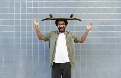 Playful man balancing skateboard on head in front of tiled wall - VRAF00490