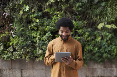 Portugal, young man using a tablet against natural plants wall background - VRAF00494