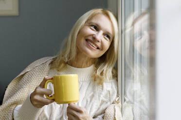 Smiling woman holding mug and looking out of window at home - TYF00815