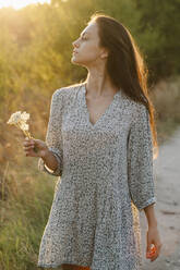 Portrait of a woman with a flower in her hands standing in a field at sunset - ALKF01112