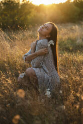 Junge Frau hockt im Gras auf einem Feld bei Sonnenuntergang - ALKF01116