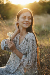 Schöne lächelnde Frau mit Blume sitzt in der Wiese bei Sonnenuntergang - ALKF01117