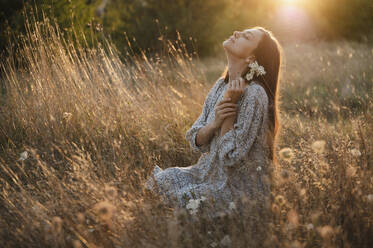 Carefree woman with flower sitting in meadow at sunset - ALKF01118