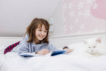 Happy girl lying on bed and reading book near cat at home - ELMF00120