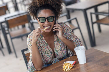 Happy woman talking on smart phone and eating french fries at sidewalk cafe - JSRF02914