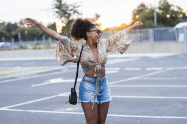 Happy woman standing with arms outstretched at parking lot - JSRF02927