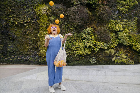 Cheerful woman throwing oranges in air standing on footpath - ALZF00004