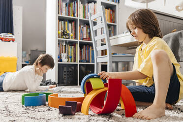 A cute preschooler boy plays with a wooden multi-colored rainbow and builds a pyramid out of it, in the background the boy is reading - ELMF00135
