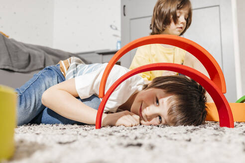 Lächelnder Junge, der auf einem Teppich liegt und durch ein gebogenes Regenbogenspielzeug mit seinem Bruder im Hintergrund schaut - ELMF00140