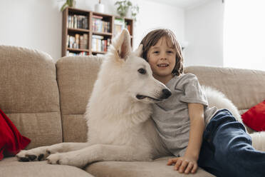Smiling boy sitting with dog on sofa at home - ELMF00144