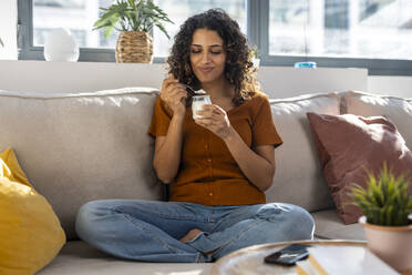 Smiling woman eating yogurt on sofa at home - JSRF02941