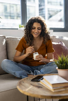 Smiling woman having yogurt on sofa at home - JSRF02943