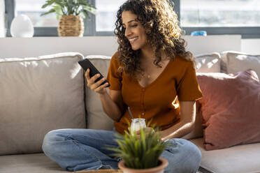 Smiling woman using smart phone on sofa at home - JSRF02946