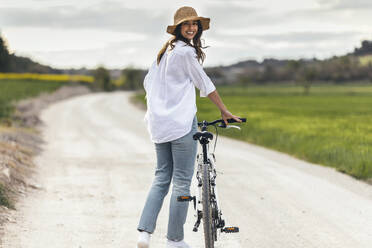 Glückliche junge Frau mit Hut und Fahrrad auf der Straße stehend - JSRF02951