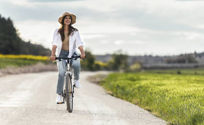 Glückliche junge Frau fährt Fahrrad auf der Straße in der Nähe eines Feldes - JSRF02954