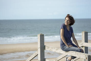 Happy woman sitting on railing at beach - KNSF10181