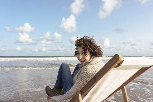 Fröhliche Frau sitzt auf einem Liegestuhl und blickt zurück auf den Strand - KNSF10196