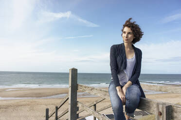 Beautiful woman looking away sitting on railing at beach - KNSF10205