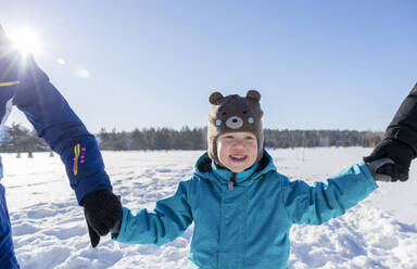 A happy boy in a blue jumpsuit holds hands with mom and dad in a field in winter. - MBLF00316