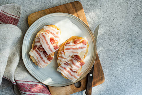 From above tow toasted bread topped with thin slices of lard, presented on a ceramic plate beside a wooden cutting board and knife. - ADSF54045