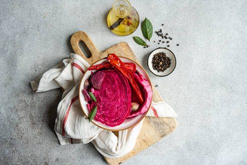 Kräftiger fermentierter Rote-Bete-Kohl mit würzigem rotem Paprika und Knoblauch, serviert in einer Schale auf einem rustikalen Holzbrett, begleitet von Olivenöl, Lorbeerblättern und ganzen Pfefferkörnern. - ADSF54054