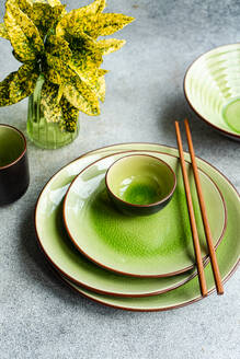 From above view of a modern table setting featuring bright green ceramic dinnerware and wooden chopsticks, juxtaposed with a vibrant potted plant, all showcased on a textured grey background. - ADSF54062