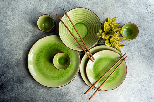 Draufsicht auf ein helles Tafelservice mit grünen Keramiktellern, Schalen und Tassen mit hölzernen Essstäbchen auf einer strukturierten Oberfläche. - ADSF54064