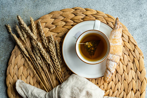 Top view of black tea with dried lemon slices in a white cup, paired with a sugar-dusted homemade croissant, placed on a woven placemat with wheat stalks. - ADSF54068