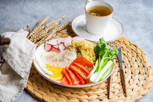 A nutritious lunch setup featuring fresh organic vegetables including reddish, tomato, and celery sticks, accompanied by a fried egg and bulgur cereal, served with a cup of fragrant tea. - ADSF54072
