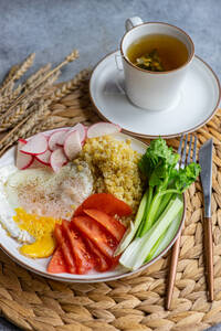 Ein appetitlicher Teller mit geschnittenem Rettich, Tomate, Sellerie, Spiegelei und Bulgur-Getreide, dazu eine Tasse heißer Tee, angerichtet auf einer Weidenmatte. - ADSF54073