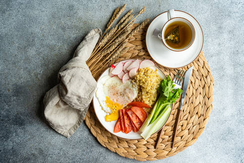 Ein ausgewogenes Mittagessen mit frischen Radieschen, Tomaten, Sellerie, einem Spiegelei und Bulgur-Getreide, begleitet von einer warmen Tasse Tee, präsentiert auf einem Tischset aus Weide. - ADSF54074