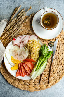 An appetizing healthy lunch set featuring a sunny side up fried egg, fluffy bulgur cereal, fresh organic radish slices, ripe tomato wedges, crisp celery sticks, and a soothing cup of tea, all artfully presented on a rustic woven placemat with wheat ears as decor. - ADSF54075