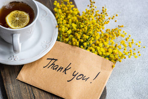 Top view of delicate cup of tea with a slice of lemon beside a cheerful bouquet of yellow Mimosa flowers and a heartfelt Thank You note - ADSF54077