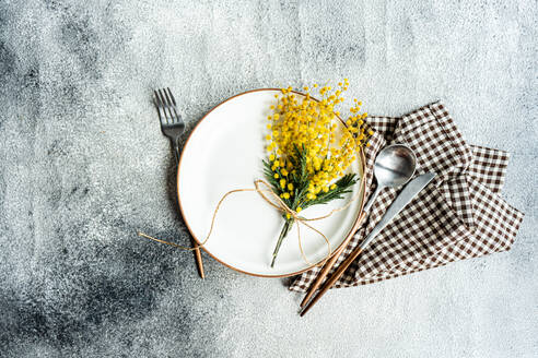 Draufsicht auf einen rustikal-schick gedeckten Tisch mit einem zarten, mit Schnur gebundenen Mimosenblumenstrauß, ergänzt durch elegantes Besteck und eine karierte Serviette - ADSF54084