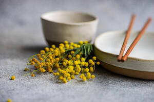 Ein ruhiger Tisch mit Keramikgeschirr, Kupferstäbchen und leuchtenden Mimosenblüten als Farbakzent - ADSF54085