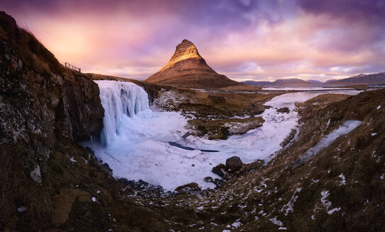 Ein Panoramablick auf einen beeindruckenden gefrorenen Wasserfall mit dem ikonischen Berg Kirkjufell unter einem dramatischen Sonnenuntergangshimmel. - ADSF54091