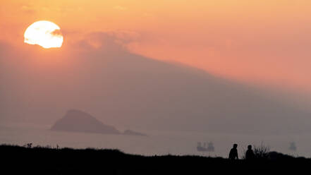 A serene seascape with a couple's silhouette against a soft orange sunset, alongside distant ships and a mountain The location is Cabo Peñas, Asturias - ADSF54150